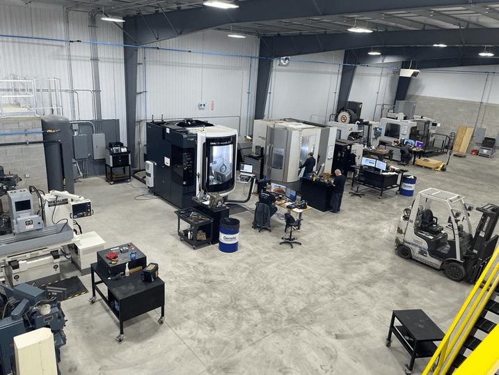 View of the machine shop from up in a corner of the warehouse wall. Along the far wall is a line of CNC mills and desks with computers for operating the machinery. The space is empty of clutter and very brightly lit.